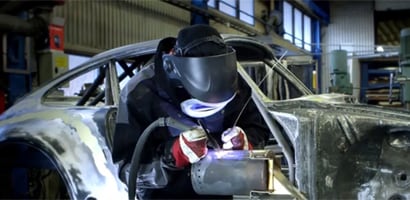 Bild på svetsare med skyddsutrustning som använder en svetspistol på en metallstruktur i en fabrik.

