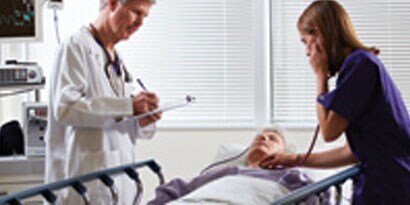 En läkare vid en patients säng, en sjuksköterska använder stetoskop på patienten
